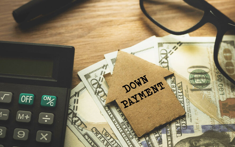 calculator, glasses, one hundred dollar bills and a small down payment sign shaped as a house on a wooden table