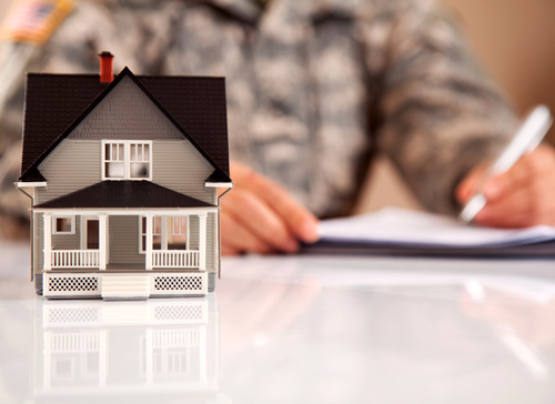 Small model house on a white surface. In the background you can see someone blurred out while signing some paperwork with a white pen