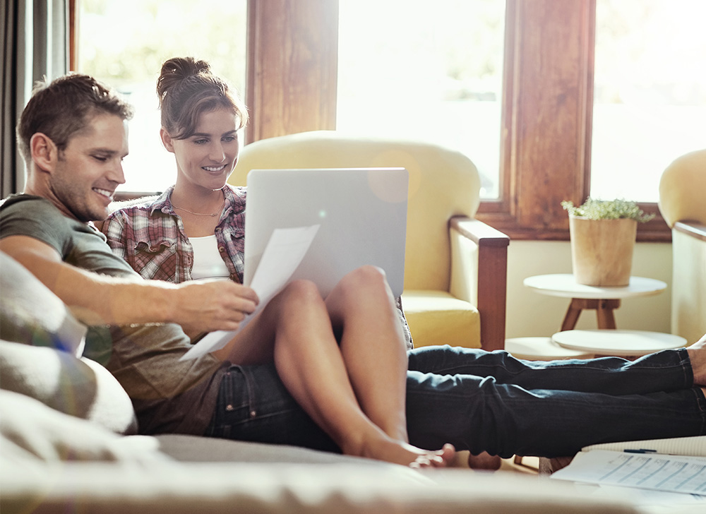 A man and a woman sitting on a couch and smiling while looking at a laptop