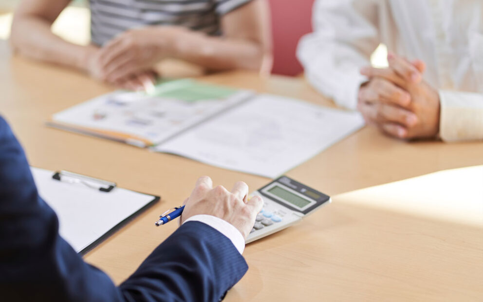 Hand of a businessman typing on a calculator in front of a couple patiently waiting.