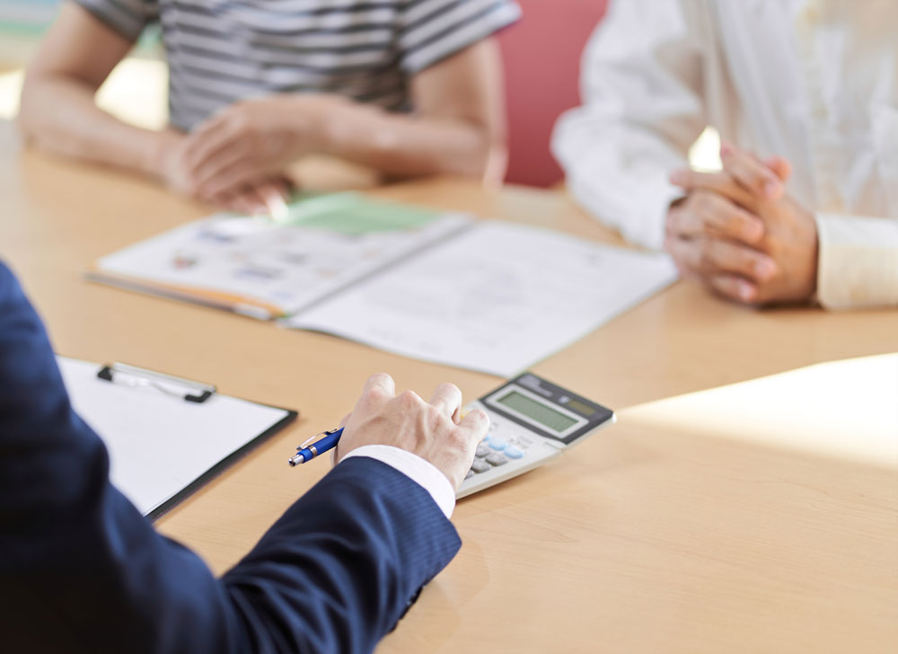 Hand of a businessman typing on a calculator in front of a couple patiently waiting.