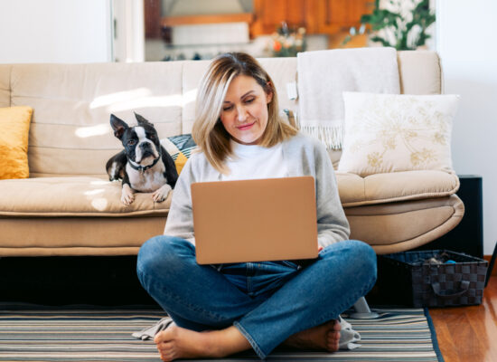 Woman working on her laptop, resting against a couch with her pet dog sitting next to her.