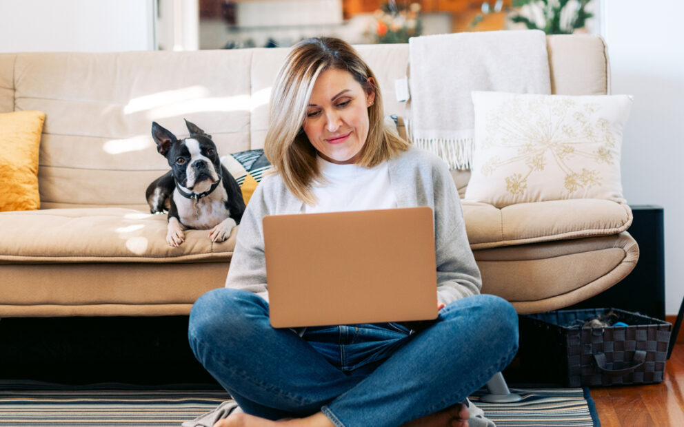Woman working on her laptop, resting against a couch with her pet dog sitting next to her.
