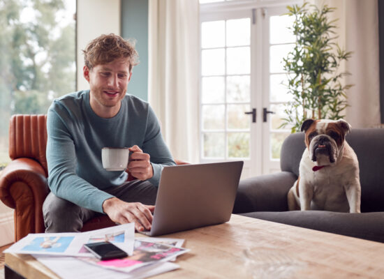 Young adult male looking at his laptop with a cup of coffee and his bulldog next to him on the couch
