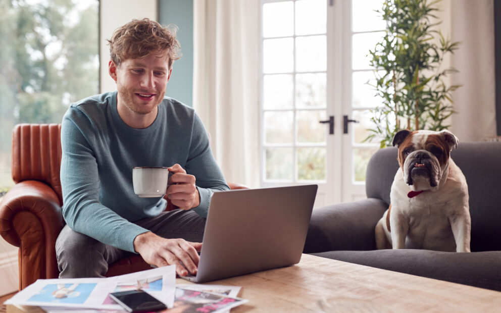 Young adult male looking at his laptop with a cup of coffee and his bulldog next to him on the couch