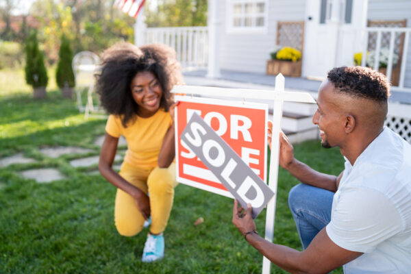 couple crounching next to a "For sale" sign, and covering it with a "sold" sign.