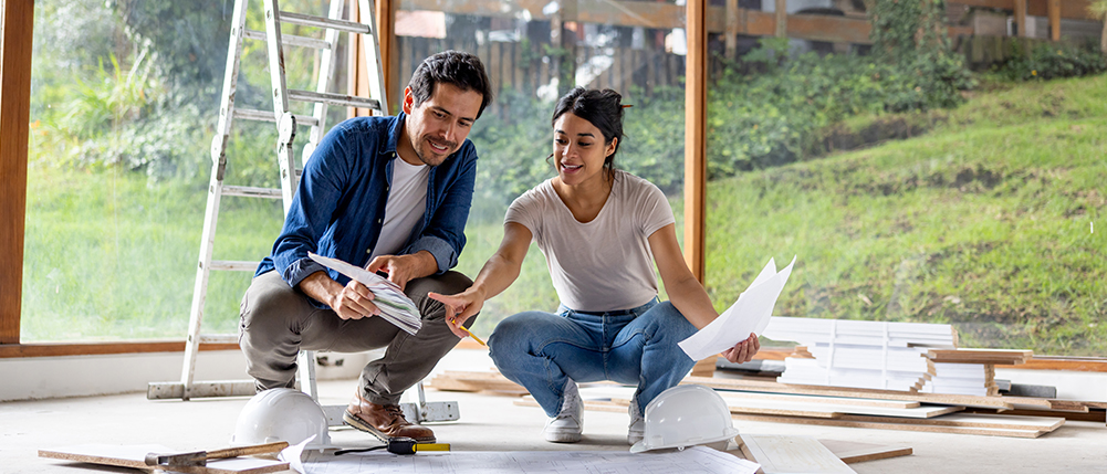 Man and woman couple looking over floor remodel plans inside room that is being renovated