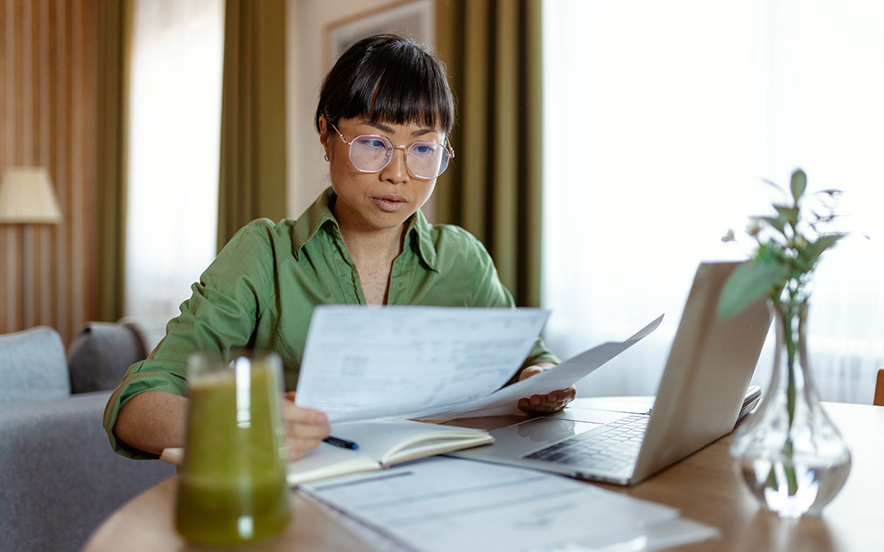 Woman sitting at home desk while comparing financial and digital documents