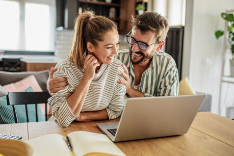 Couple smiling while sitting at kitchen table and looking at laptop