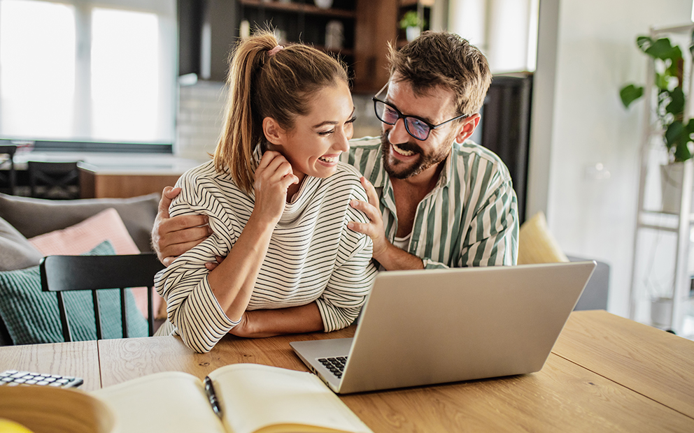 Couple smiling while sitting at kitchen table and looking at laptop