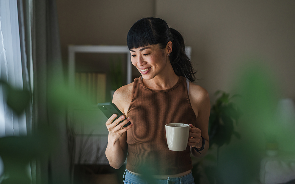 Woman drinking coffee at home while checking her cell phone