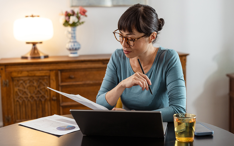 Middle aged woman sitting at home desk reviewing information on paper and her laptop