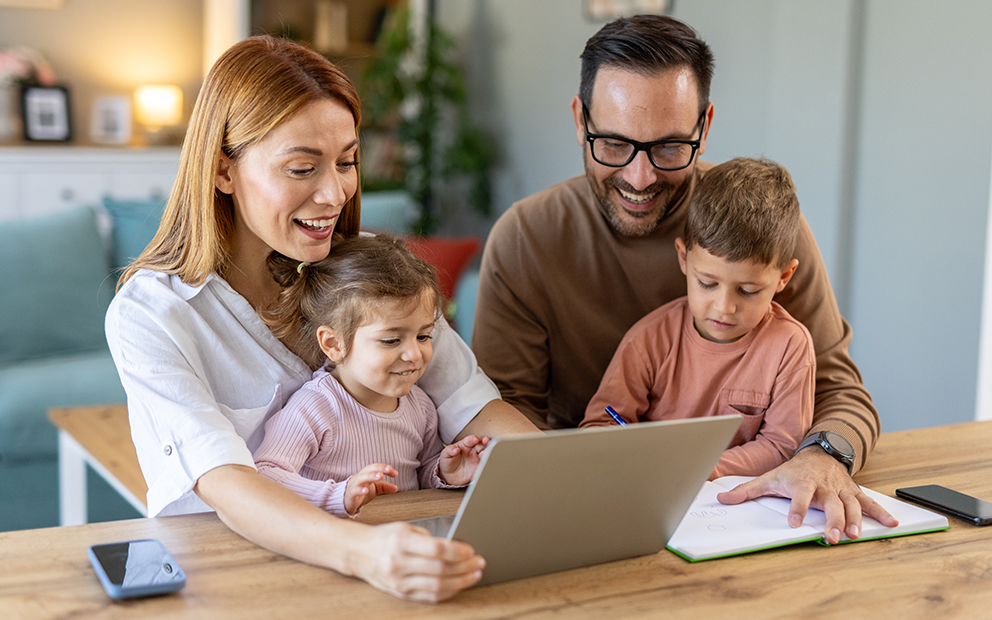 Mom and dad sitting at table looking at laptop with their own children sitting in their laps