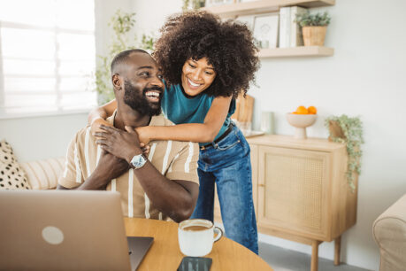 Woman hugging male partner from behind while he sits at desk looking at computer