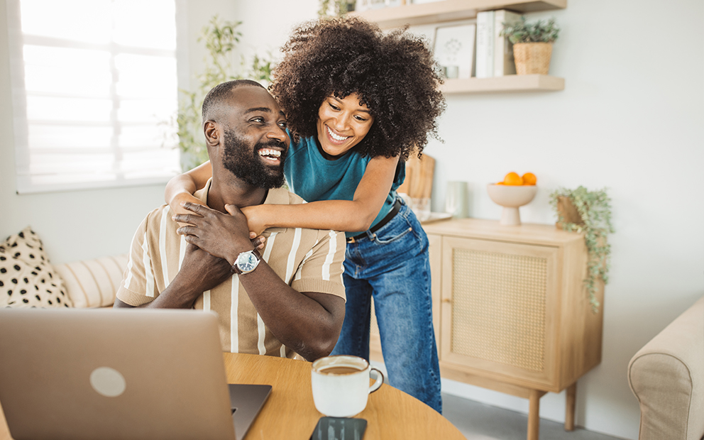 Woman hugging male partner from behind while he sits at desk looking at computer