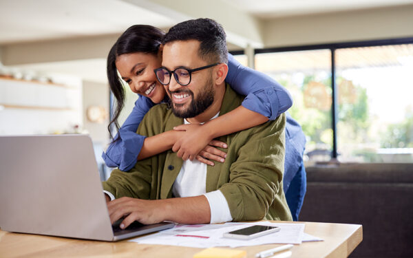Woman hugging male partner from behind while he sits at desk looking at computer